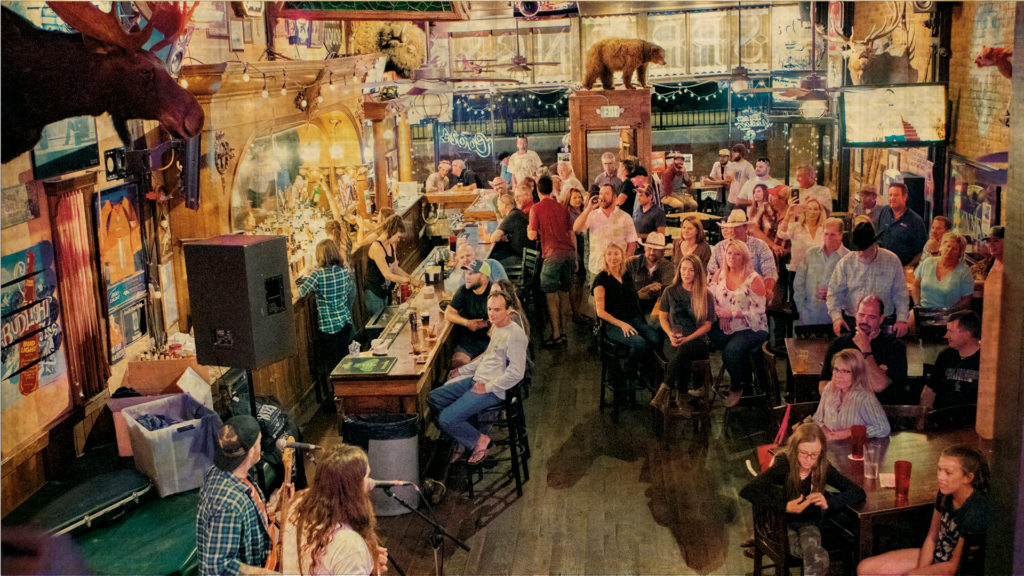 A shot for high up in a western bar adorned with stuffed animal heads, and occupied with a large crowd of happy people eating and drinking while they listen to a band that is situated at the front of the image